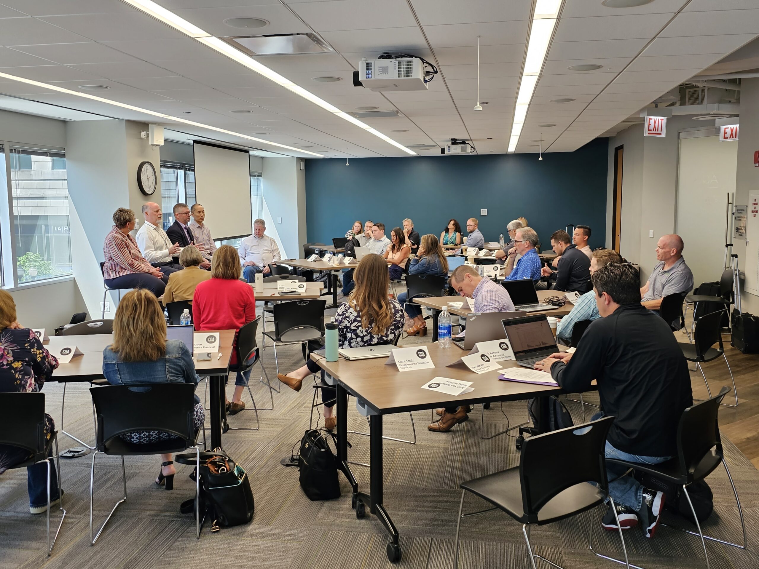 A panel discussion in a modern meeting room, with attendees seated around tables engaging in discussion.