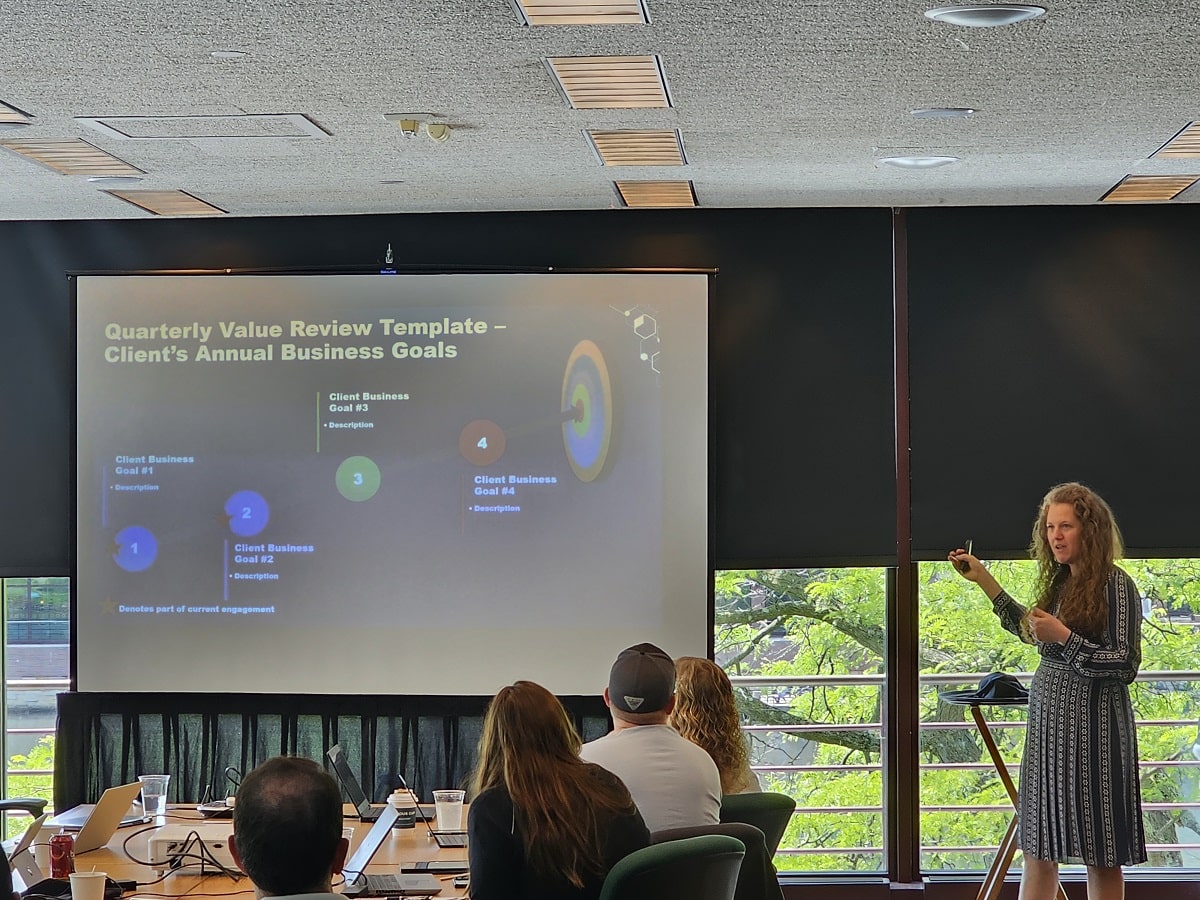 A woman presenting a slide on "Quarterly Value Review Template – Client’s Annual Business Goals" to an engaged audience.