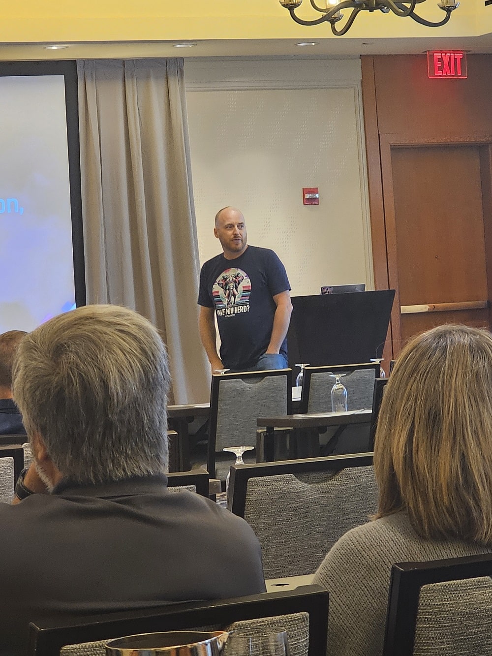 A man in a casual t-shirt presenting in a conference room while attendees listen attentively.