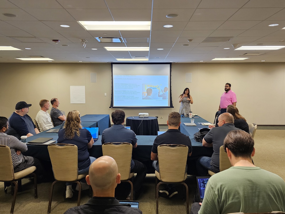 A professional conference room with two presenters, a woman and a man, leading a discussion while attendees listen.